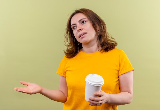 Young casual woman holding plastic coffee cup and showing empty hand looking at left side on isolated green wall