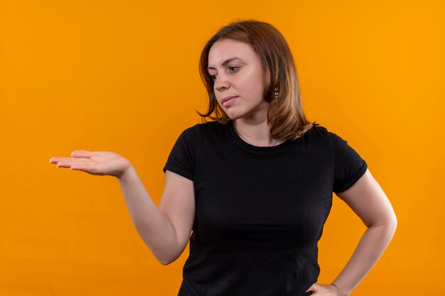 Young casual woman looking at left side and showing empty hand on isolated orange wall