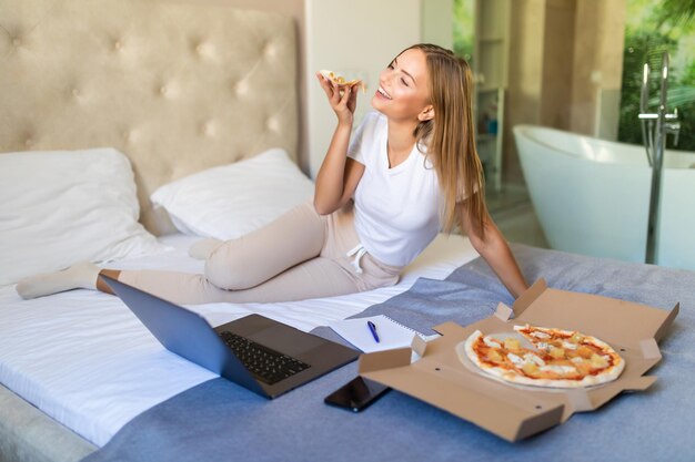 ピザを食べてラップトップを使用しながら自宅のベッドでベッドに横になっている若いカジュアルな女性
