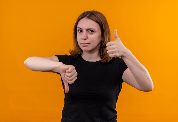 Молодая случайная женщина показывает палец вверх и вниз, глядя на изолированную оранжевую стену