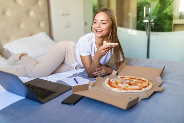 Young casual woman with pizza using laptop while resting on bed at home.