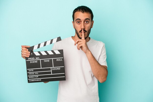 Young caucasian actor man holding clapperboard isolated on blue background keeping a secret or asking for silence.