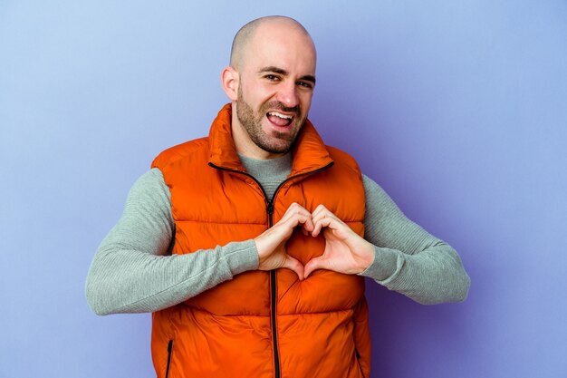 Young caucasian bald man on purple smiling and showing a heart shape with hands.