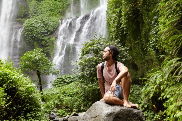 Молодой кавказский босоногий турист с рюкзаком сидит на скале в окружении тропических лесов и любуется великолепным видом на водопад