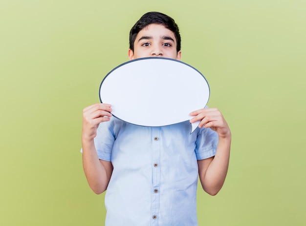 Young caucasian boy holding speech bubble looking at camera from behind it isolated on olive green background with copy space