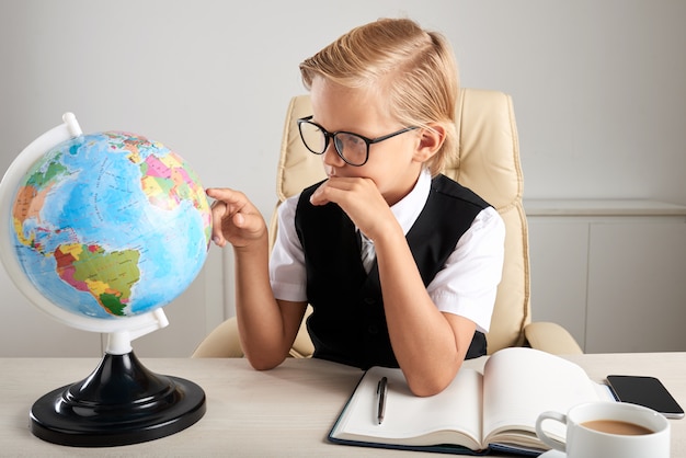 Free photo young caucasian boy sitting in executive chair in office and looking at earth globe