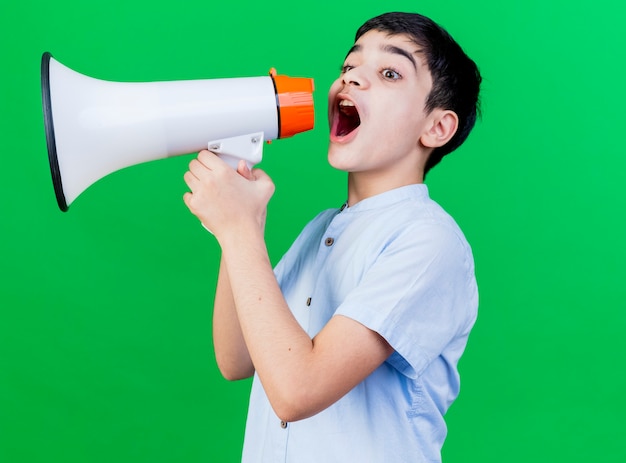 Young caucasian boy standing in profile view talking by speaker isolated on green background with copy space