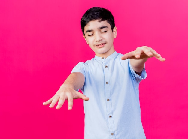 Free photo young caucasian boy walking with closed eyes stretching out hands towards camera isolated on crimson background