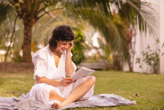 Young caucasian brunette girl in white dress uses drawing papers sitting crosslegged on grass Summer holiday concept