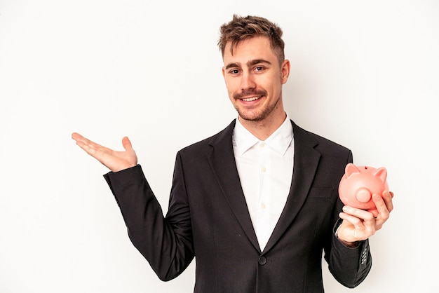 Young caucasian business man holding piggy bank isolated on white background showing a copy space on a palm and holding another hand on waist.
