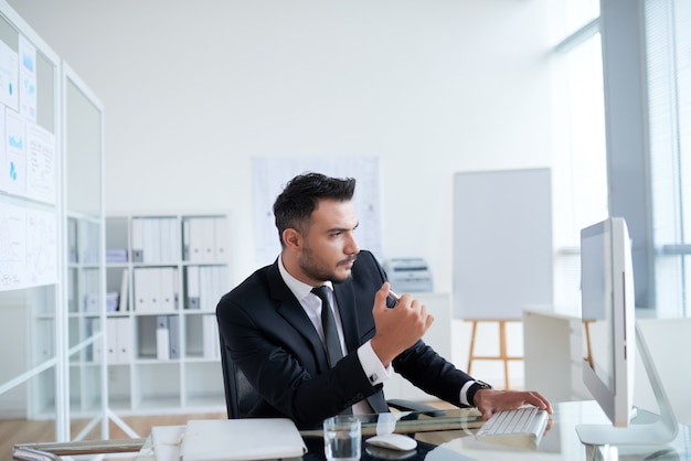 Free photo young caucasian businessman in suit sitting in office and looking at computer screen