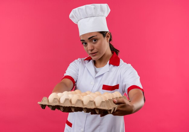 Young caucasian cook girl in chef uniform holds batch of eggs out isolated on pink background with copy space