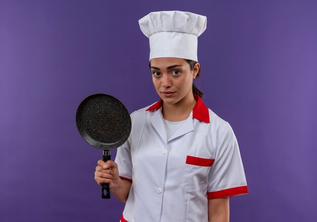 Young caucasian cook girl in chef uniform holds frying pan and looks at camera isolated on violet background with copy space