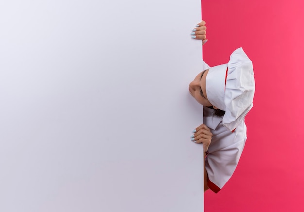Young caucasian cook girl in chef uniform stands behind and looks at white wall on pink  with copy space