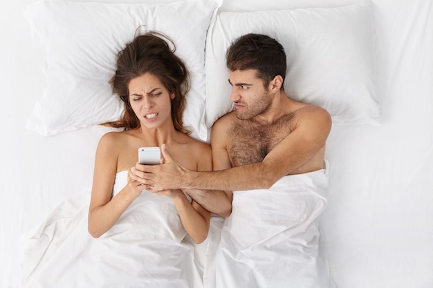 ベッドで戦っている若い白人カップル：ひげを剃っていない男が妻の手から携帯電話をひったくろう