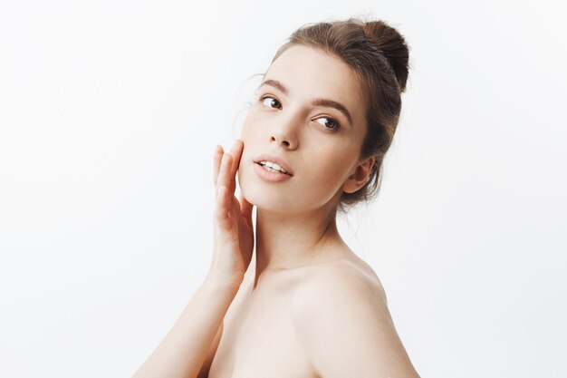 young caucasian dark-haired student girl with bun hairstyle and naked body touching skin on face with fingers, lookin aside with calm and relaxed face expression.