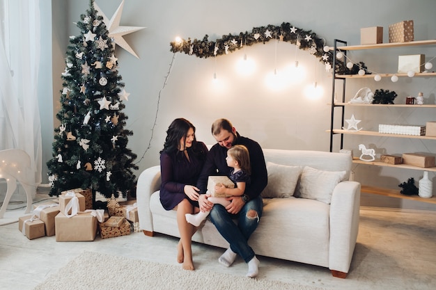 Young caucasian family of three sit on the sofa next to the Christmas tree together