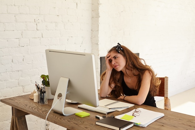 Young Caucasian female designer feels panic from the coming deadline for her work, sitting at workplace with papers, notepad and looks in the computer monitor against white brick wall, looking puzzled