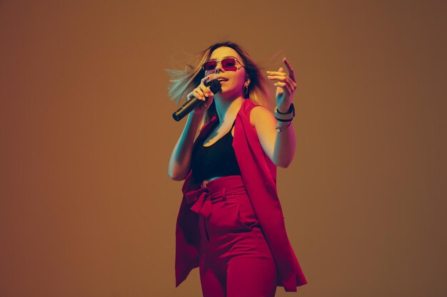 Young caucasian female musician, performer singing, dancing in neon light on gradient