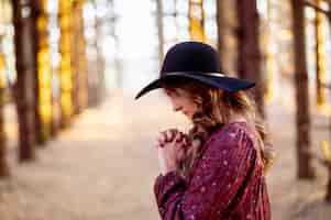 Foto gratuita giovane femmina caucasica in un cappello nero alla moda che prega nella foresta scenica, umore di autunno