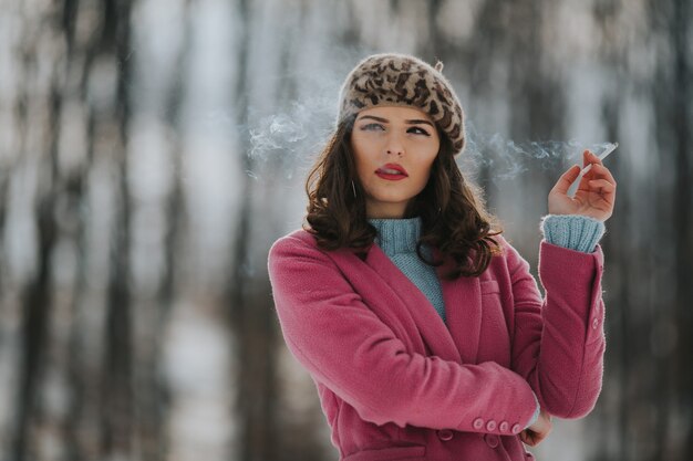 ピンクのコートを着て、背景に木がある公園で喫煙している若い白人女性