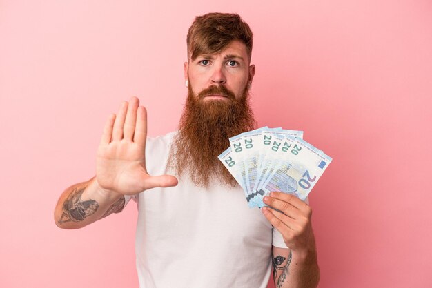 ピンクの背景に分離された紙幣を保持している長いひげを持つ若い白人生姜男は、一時停止の標識を示す伸ばした手で立って、あなたを防ぎます。
