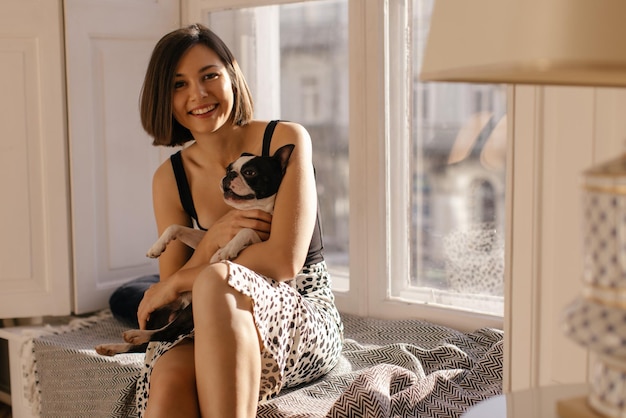 Free photo young caucasian girl holding dog and smiling looking at camera while sitting on windowsill pets concept