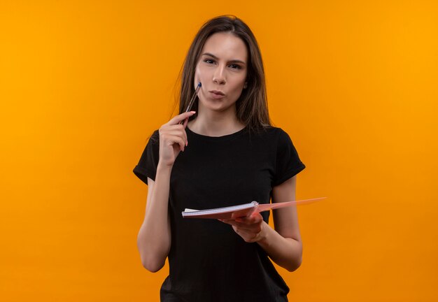 young caucasian girl wearing black t-shirt holding notebook put pen on cheek on isolated orange wall