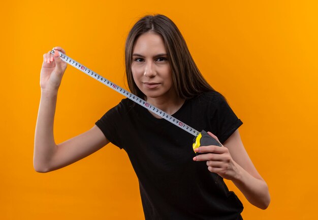 young caucasian girl wearing black t-shirt holding tape measure on isolated orange wall