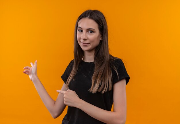 young caucasian girl wearing black t-shirt points at back on isolated orange wall