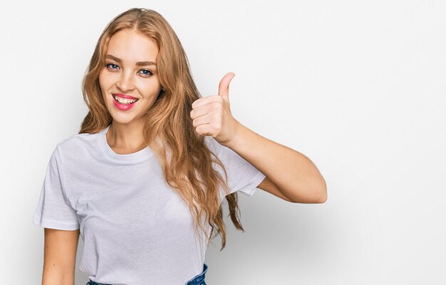Молодая кавказская девушка в повседневной белой футболке улыбается счастливой и позитивной, показывает большой палец вверх, делает отлично и знак одобрения