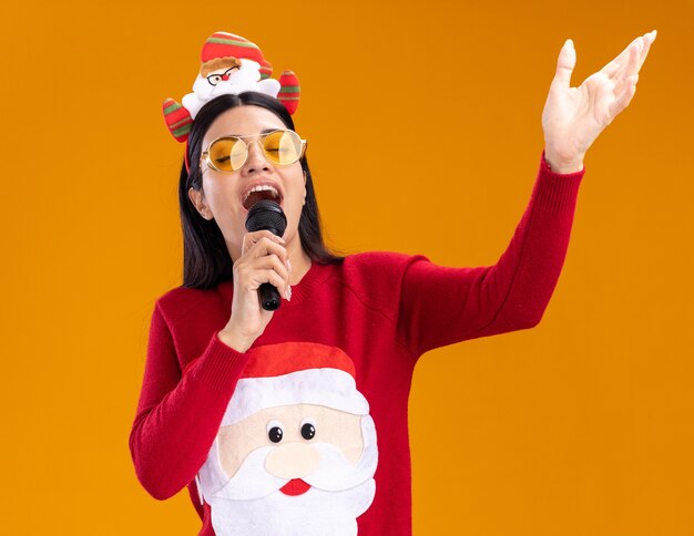 サンタクロースのヘッドバンドとセーターを着て、口の近くでマイクを持って、オレンジ色の背景で隔離された空気の中で手を閉じて歌っている白人の少女