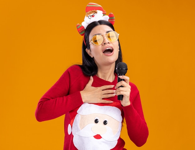 молодая кавказская девушка в головной повязке санта-клауса и свитере в очках держит микрофон возле рта поет с закрытыми глазами, держа руку на груди, изолированной на оранжевой стене с копией пространства