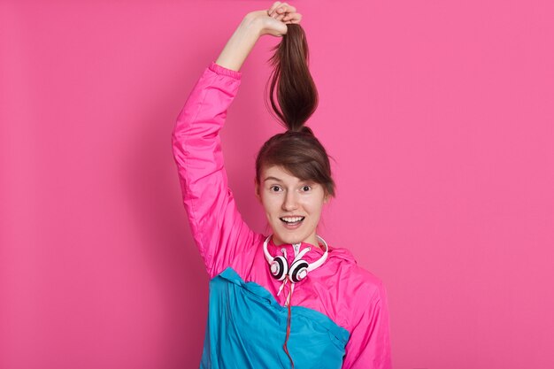 young Caucasian girl with dark hair holding her ponitail by hands playing with it, smiles, keeps mouth opened, lady with pleasant appearance posing isolated on pink.