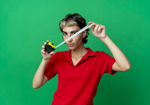 Young caucasian girl with pixie haircut holding tape meter looking at camera isolated on green background with copy space