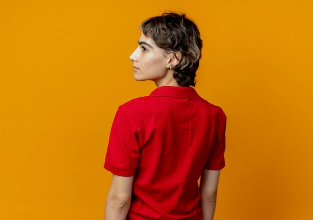 Young caucasian girl with pixie haircut standing in behind view looking at side isolated on orange background with copy space