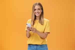 Бесплатное фото Молодая кавказская девушка с загорелой кожей и светлыми волосами использует беспроводные наушники, чтобы позвонить другу через смартфон, прижав мобильный телефон к груди, весело улыбаясь в камеру, привыкая к новым технологиям