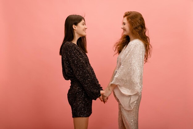 Молодые кавказские девушки в блестящих платьях улыбаются, смотрят друг на друга и держатся за руки на розовом фоне Концепция женской дружбы