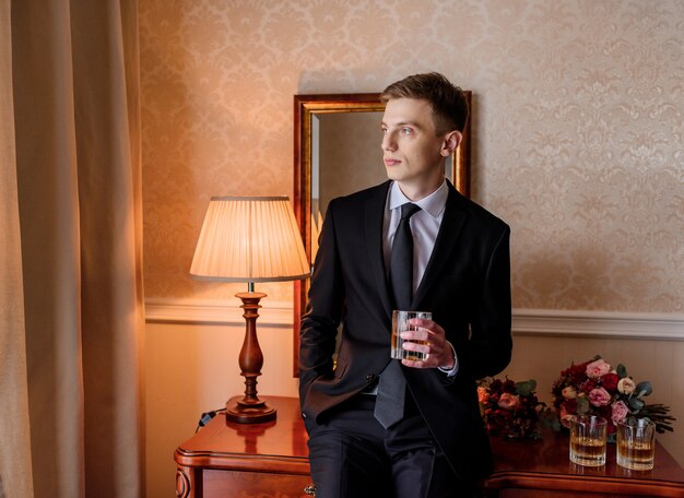 Young caucasian groom dressed in stylish tuxedo drinking alcohol in room and sitting on the table next to bridal bouquets