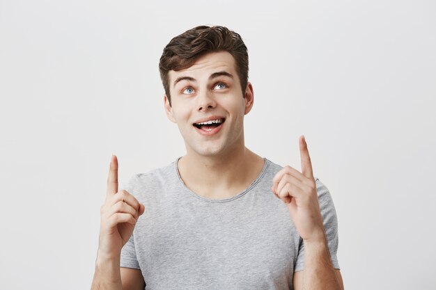 Young caucasian guy wearing gray t-shirt pointing at copy space up with forefingers looking with broad smile upwards demonstrating something at white studio background. Body language concept