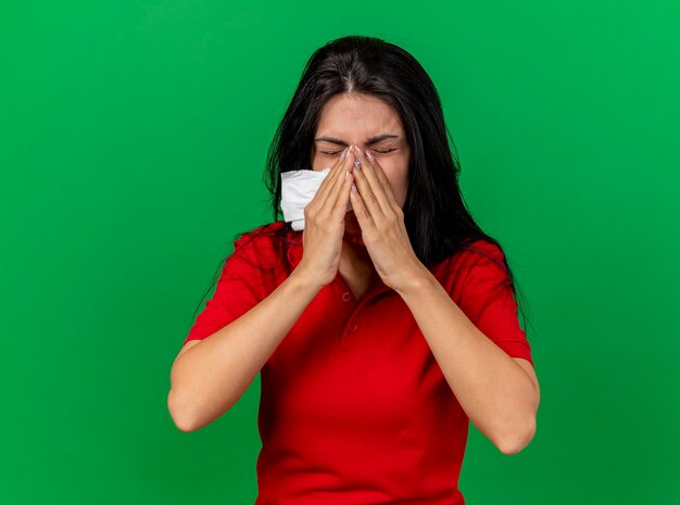 Young caucasian ill girl holding napkin keeping hands on nose sneezing isolated on green wall with copy space