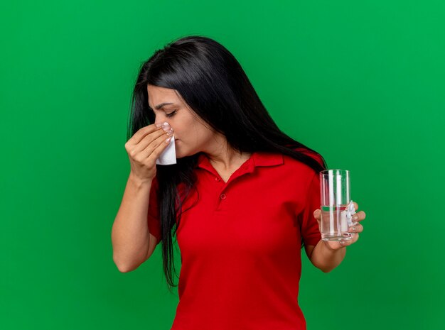 Young caucasian ill girl holding pack of tablets glass of water and wiping nose with napkin with closed eyes isolated on green background with copy space