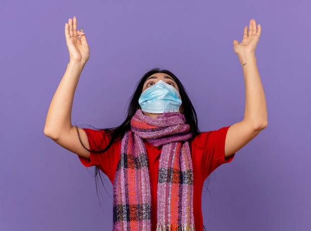 Young caucasian ill girl wearing mask and scarf holding thermometer looking up and raising hands up praying and blessing god isolated on purple wall