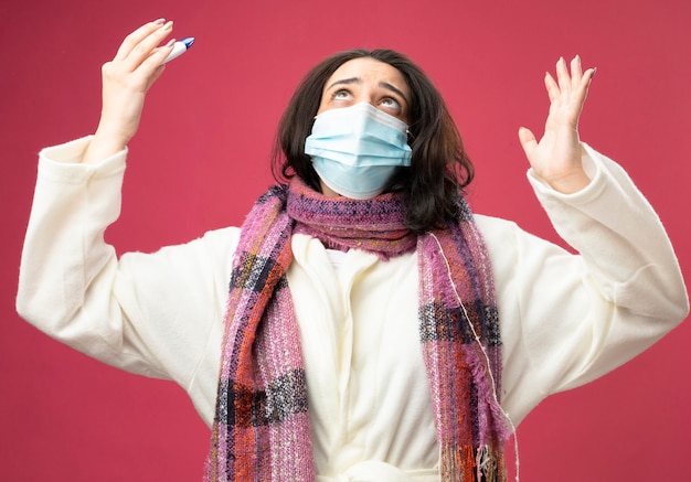 Foto gratuita giovane ragazza malata caucasica che indossa veste e sciarpa con maschera che tiene termometro alzando la mano alzando lo sguardo pregando e benedicendo dio isolato sulla parete cremisi