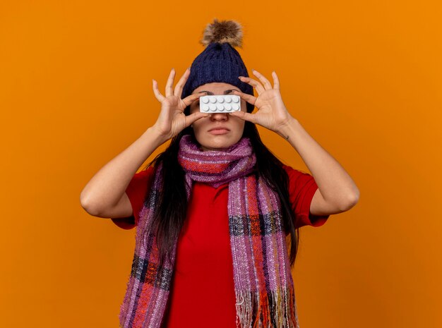 Young caucasian ill girl wearing winter hat and scarf holding pack of tablets in front of eyes isolated on orange wall
