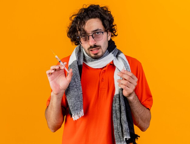 Young caucasian ill man wearing glasses and scarf holding syringe and ampoule looking at camera isolated on orange background