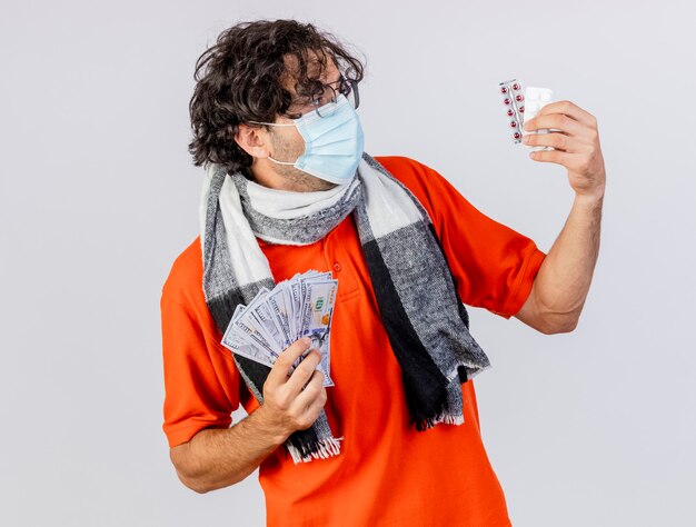 白い背景で隔離の医療ピルを見てお金と医療ピルを保持しているメガネスカーフとマスクを身に着けている若い白人の病気の男