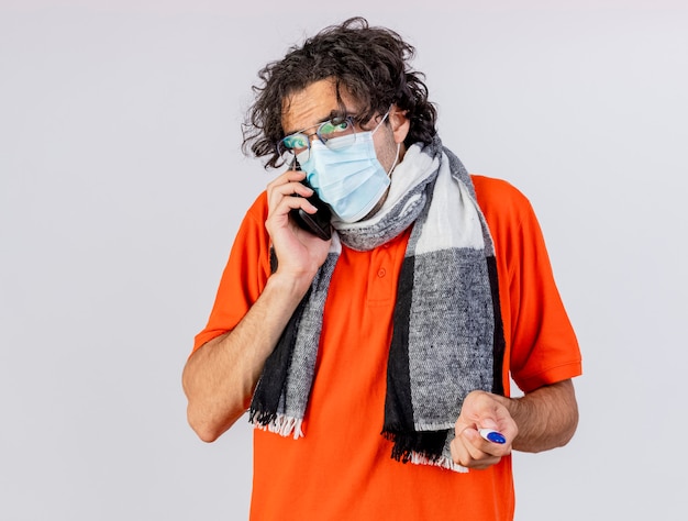 Молодой кавказский больной человек в очках, шарфе и маске, держащий термометр, разговаривает по телефону, изолированном на белой стене с копией пространства