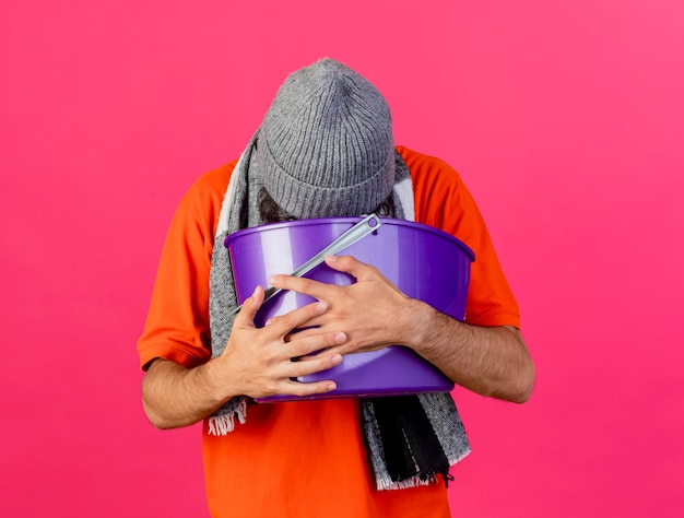 Free photo young caucasian ill man wearing glasses winter hat and scarf holding plastic bucket having nausea vomiting into bucket isolated on crimson wall