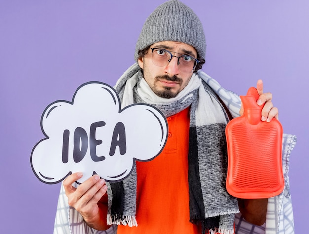 Young caucasian ill man wearing glasses winter hat and scarf wrapped in plaid holding idea bubble and hot water bag looking at camera isolated on purple background